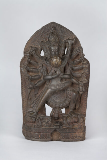 Earth-colored relief sculpture depicting many-armed deity. standing atop vanquished foes, in embrace with consort