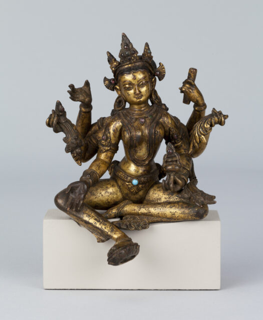 Tarnished golden statue depicting six-armed goddess wearing pointed crown holding implements in each hand