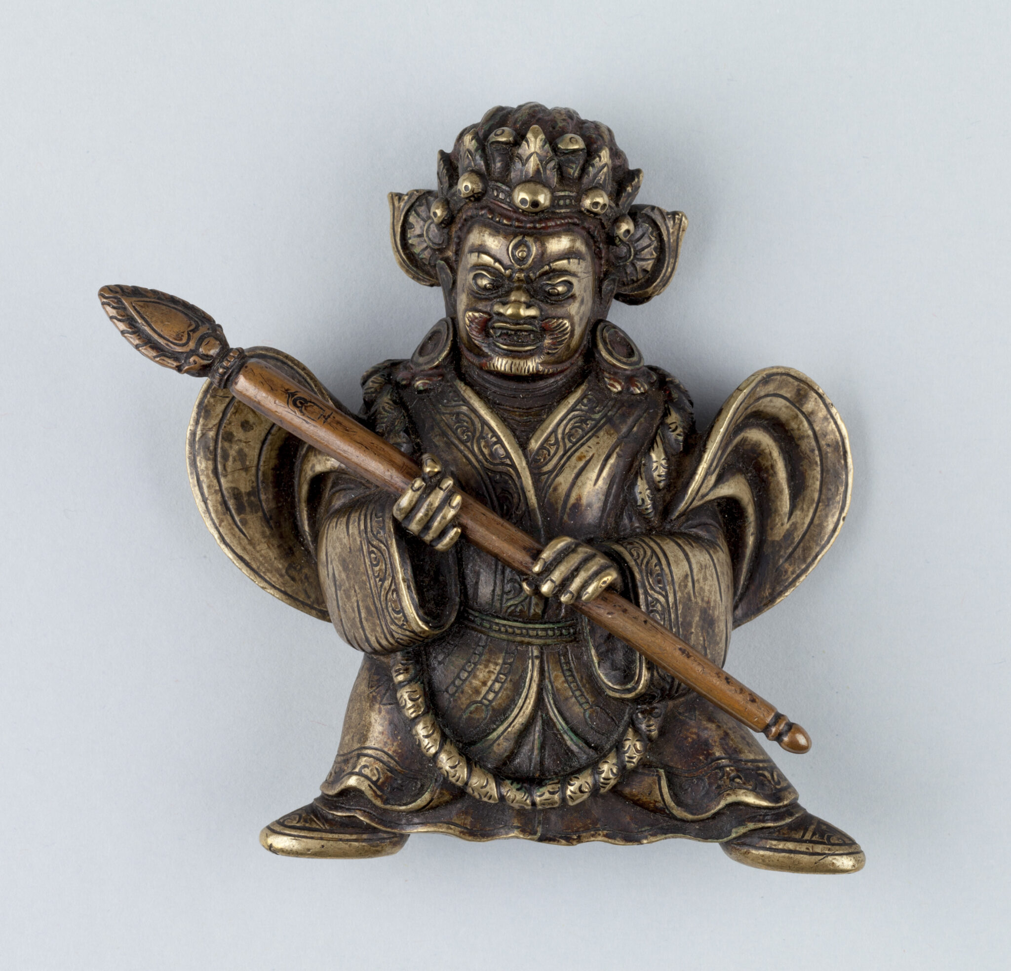 Tarnished brass-colored sculpture depicting wrathful deity wearing crown and billowing robes holding staff across body