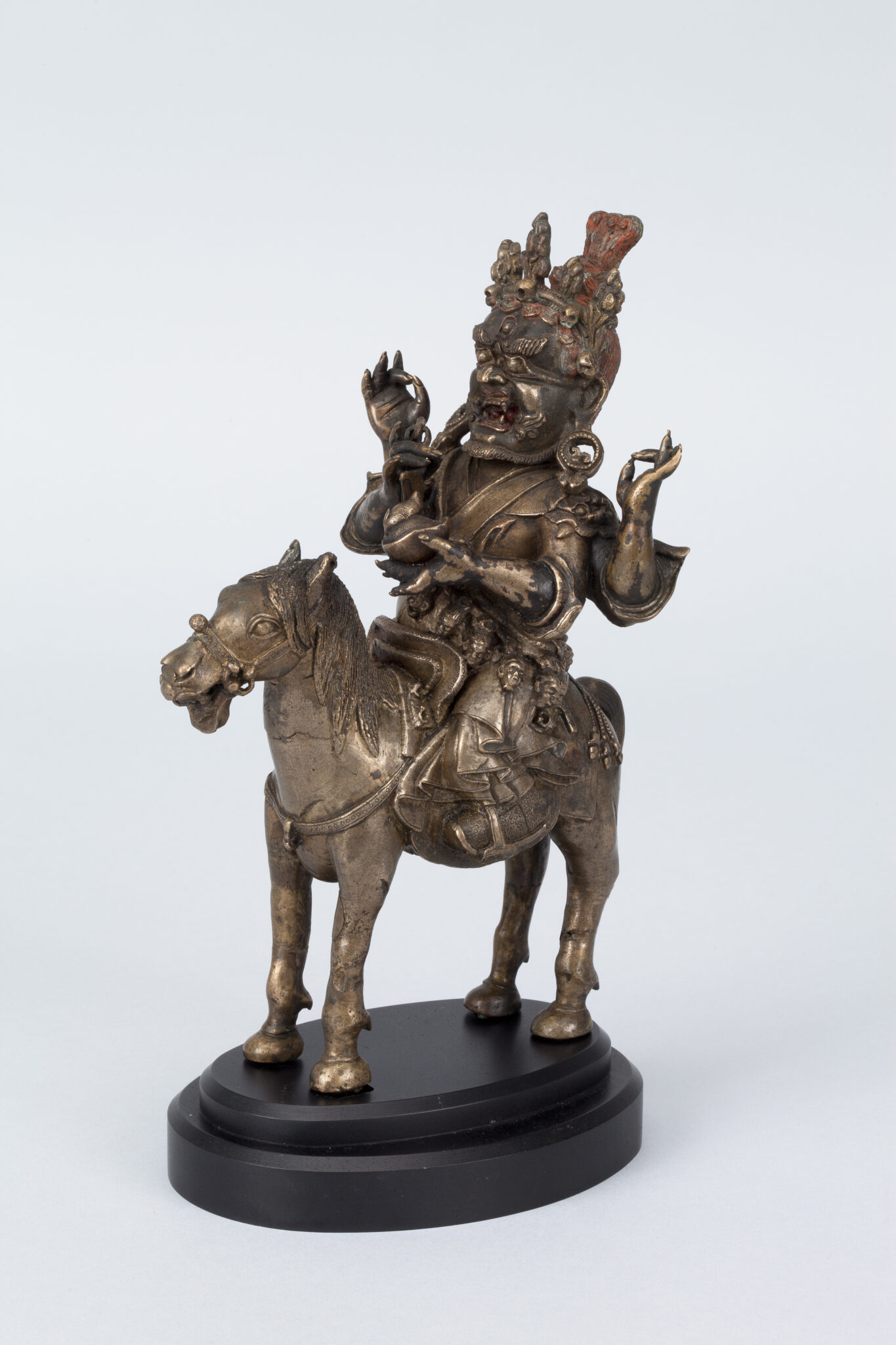 Brass-colored sculpture depicting four-armed wrathful deity with mouth open in grimace riding atop horse
