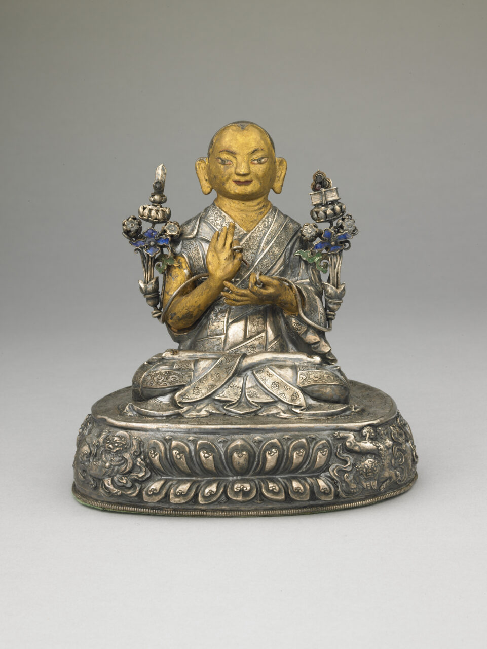 Gold- and silver-colored sculpture depicting monk wearing pleated robe, holding long-stemmed blossoms in hands