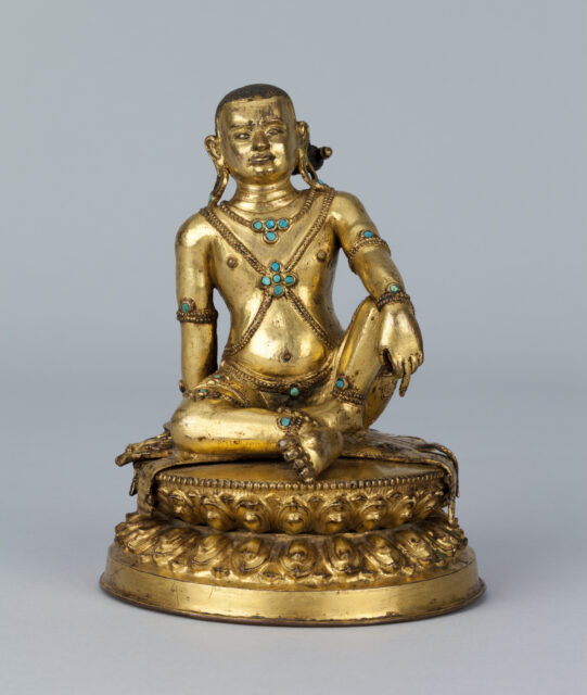 Golden sculpture depicting bare-chested figure, seated with right knee raised, wearing turquoise-accented jewelry