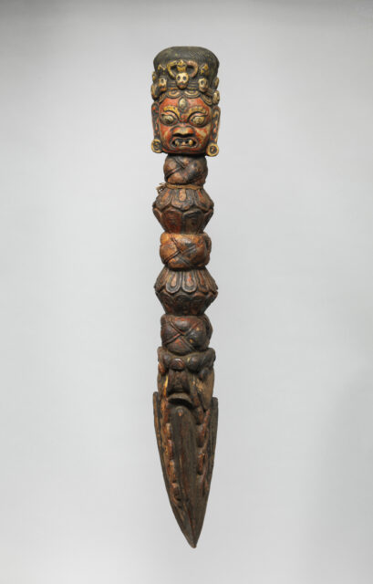 Carved wooden dagger-shaped implement featuring head, face scowling, wearing crown of skulls at pommel