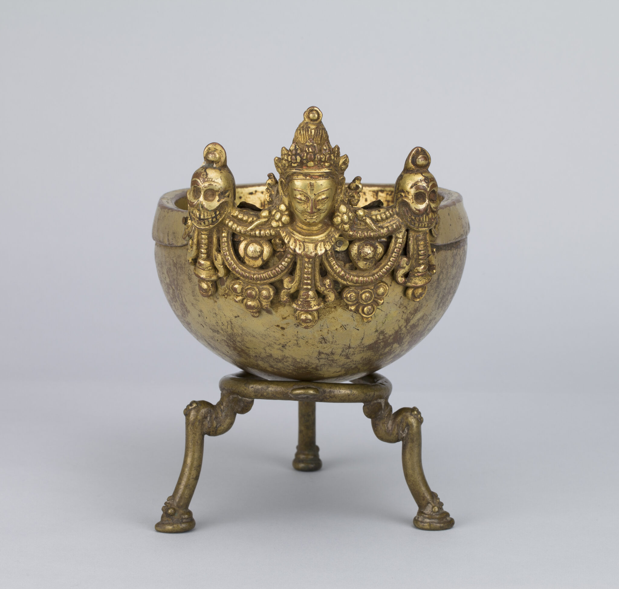 Cup made from human skull mounted on brass tripod, decorated with face of deity, skulls, and garland motif