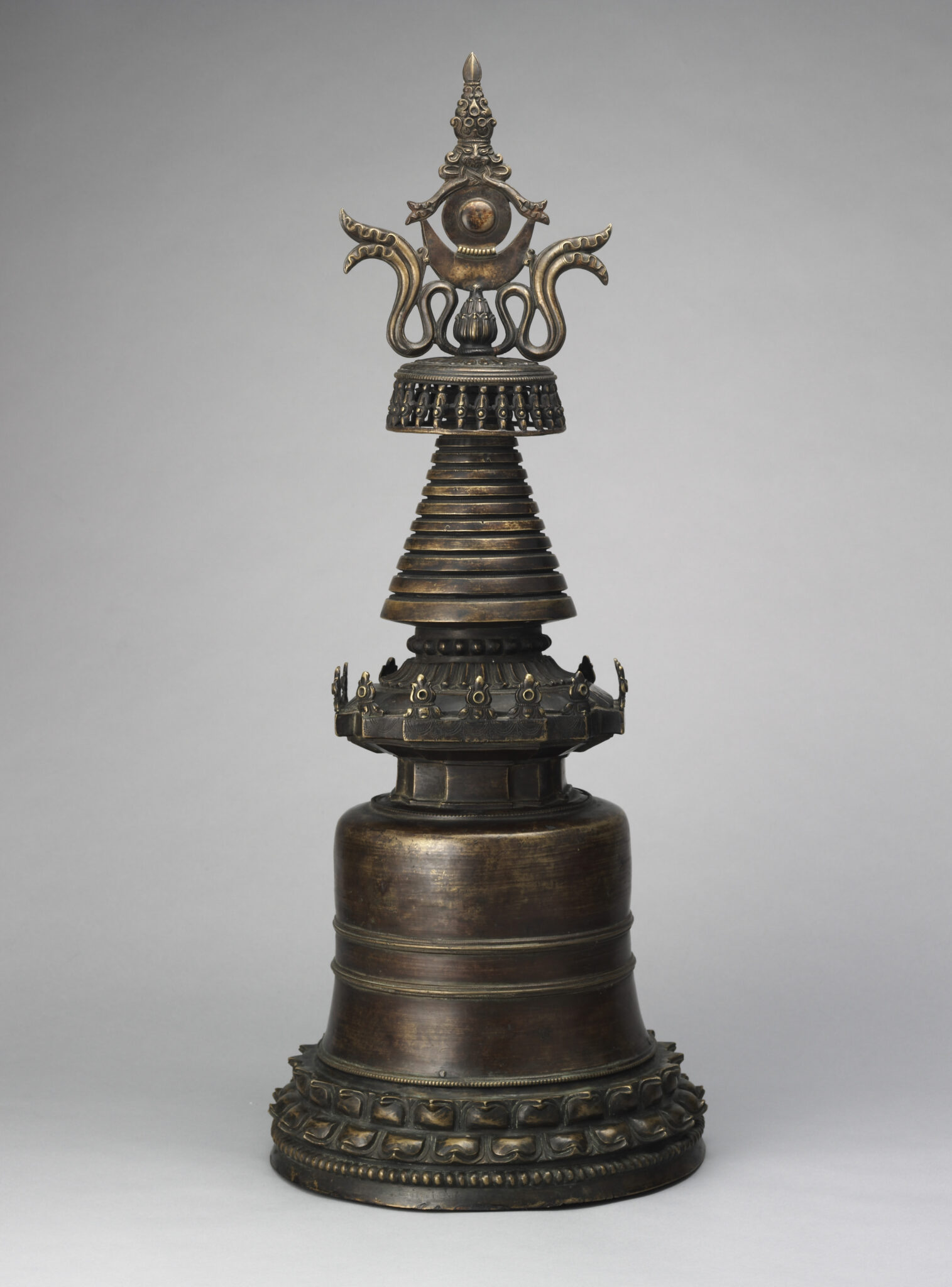 Patinated metal religious implement in shape of stupa resting on lotus pedestal featuring serpentine motif at apex