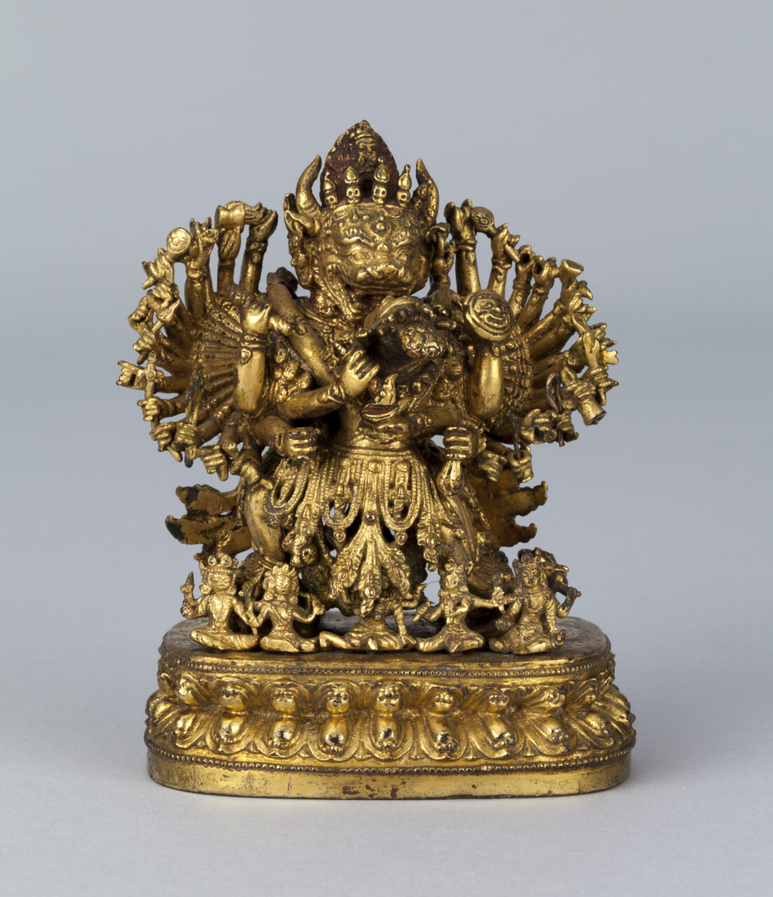 Golden sculpture depicting cow-headed, many-armed deity in embrace with consort surrounded by circle of deities