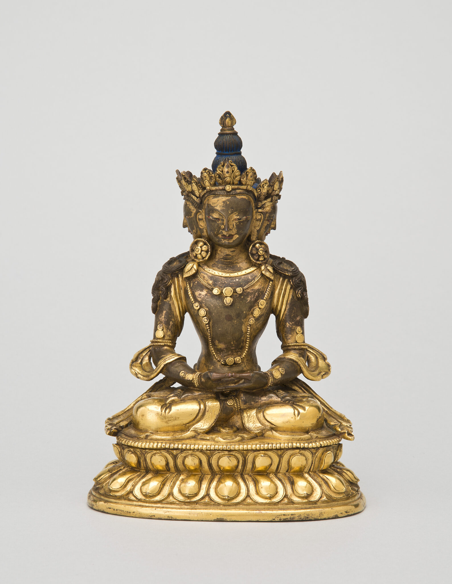 Patinated golden sculpture depicting Buddha wearing pointed crown and tall hairstyle seated in meditation