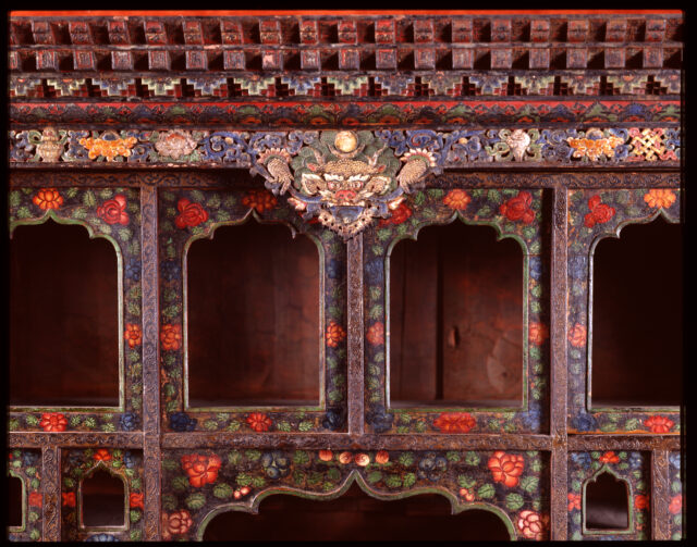 Detail of top-middle of cabinet: Dragon’s head situated at center of furniture; niche frames painted abundantly with blossoms
