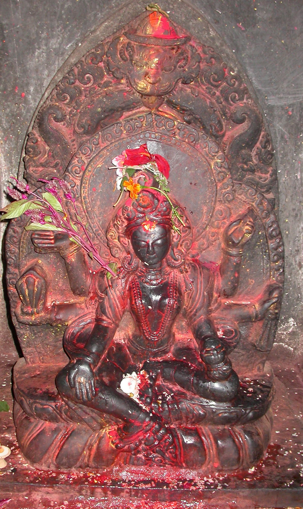 Dark stone sculpture of six-armed deity anointed with offerings of pigment and flowers