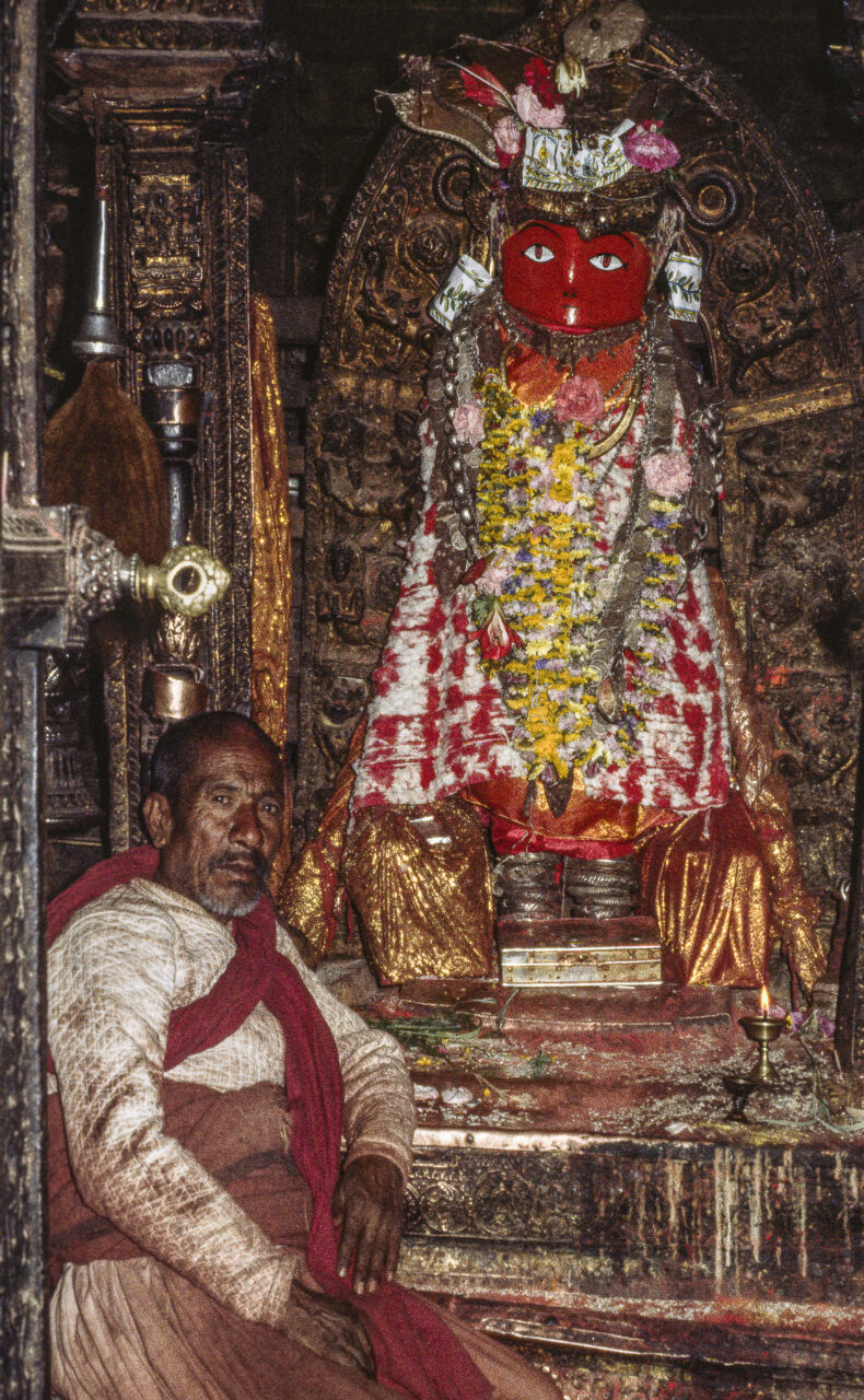 Man sits before statue adorned with garlands and textiles; statue has red, abstract face
