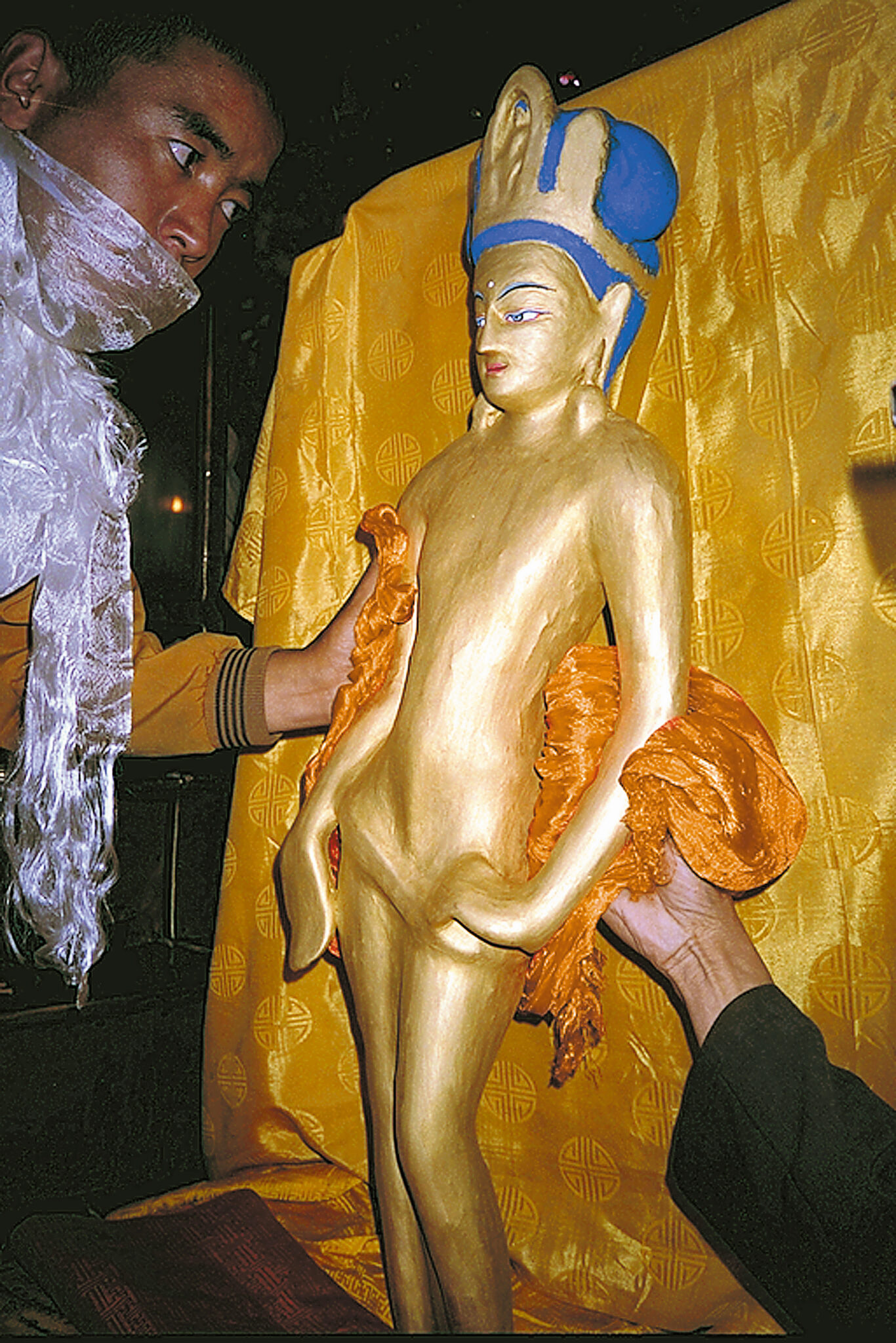 Golden statue of crowned deity with blue hair handled delicately by people with saffron-colored textiles