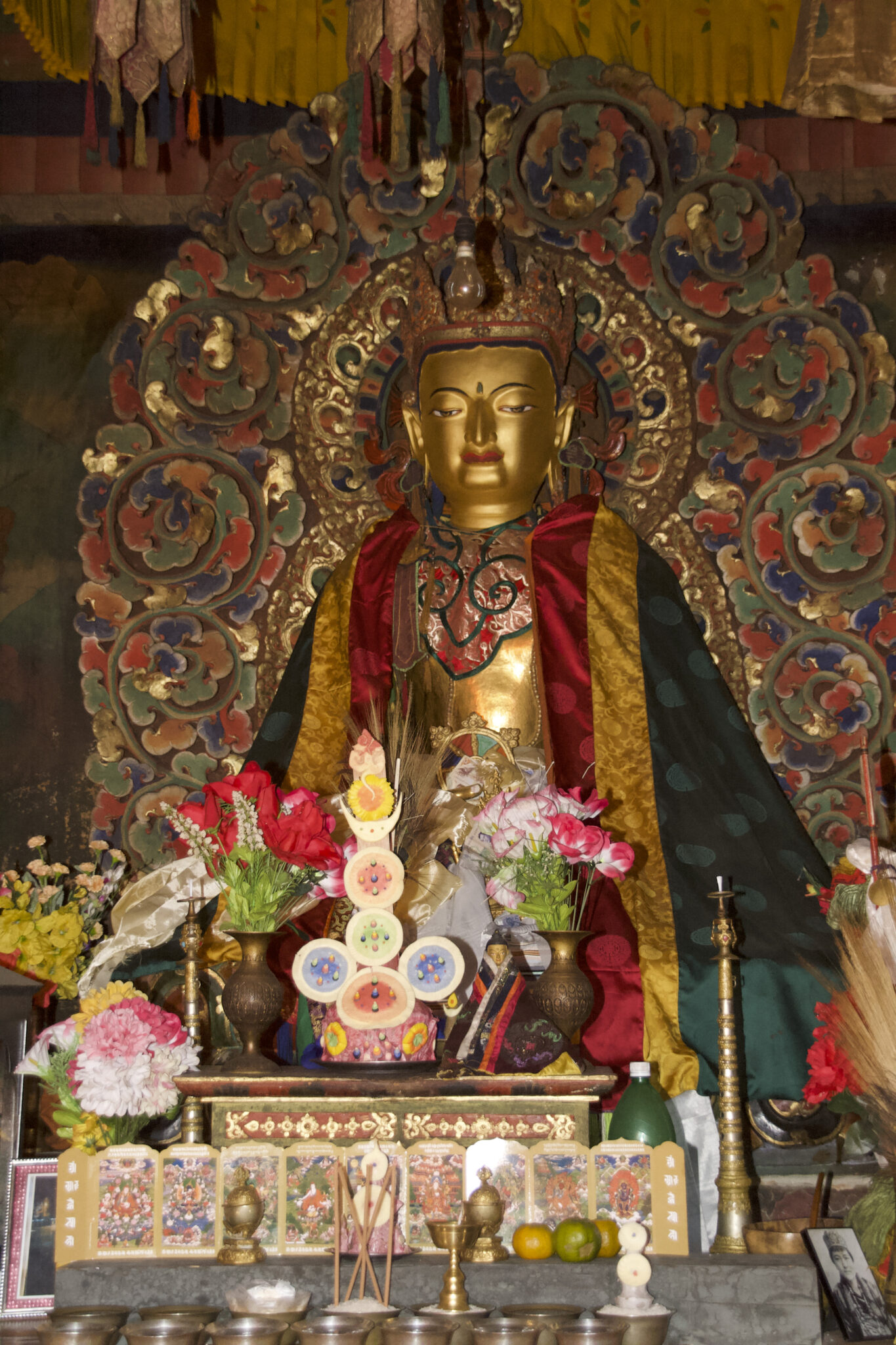 Golden Buddha dressed in green, saffron, and red cape seated between altar and colorful mandorla