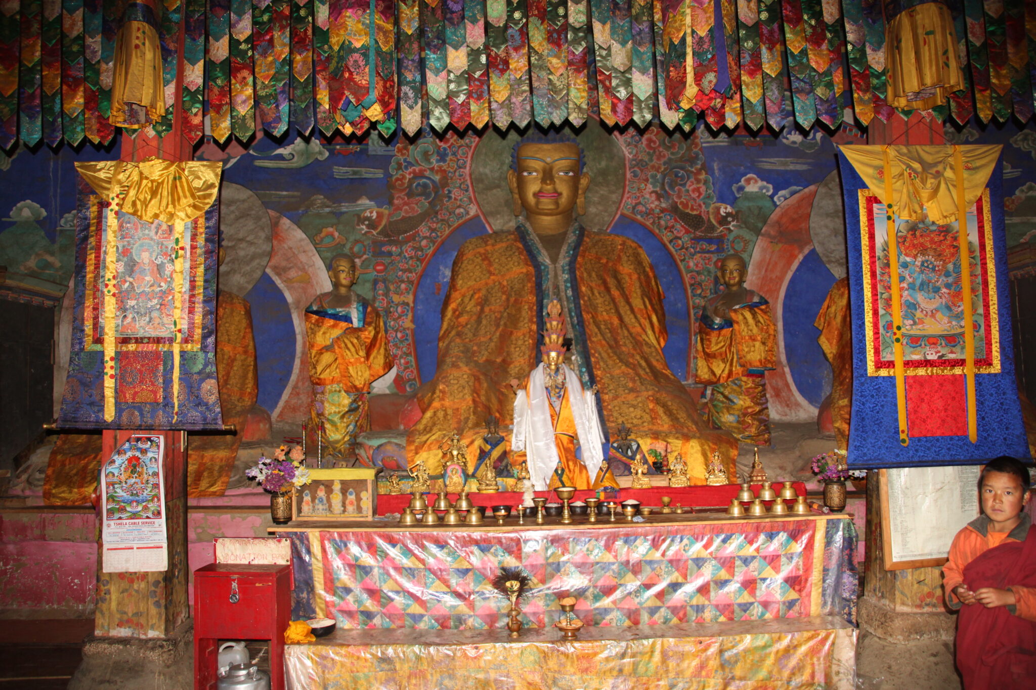 Buddha wearing saffron robe seated in niche underneath colorful banners and between two thangkas (paintings on cloth with embroidered textile borders)