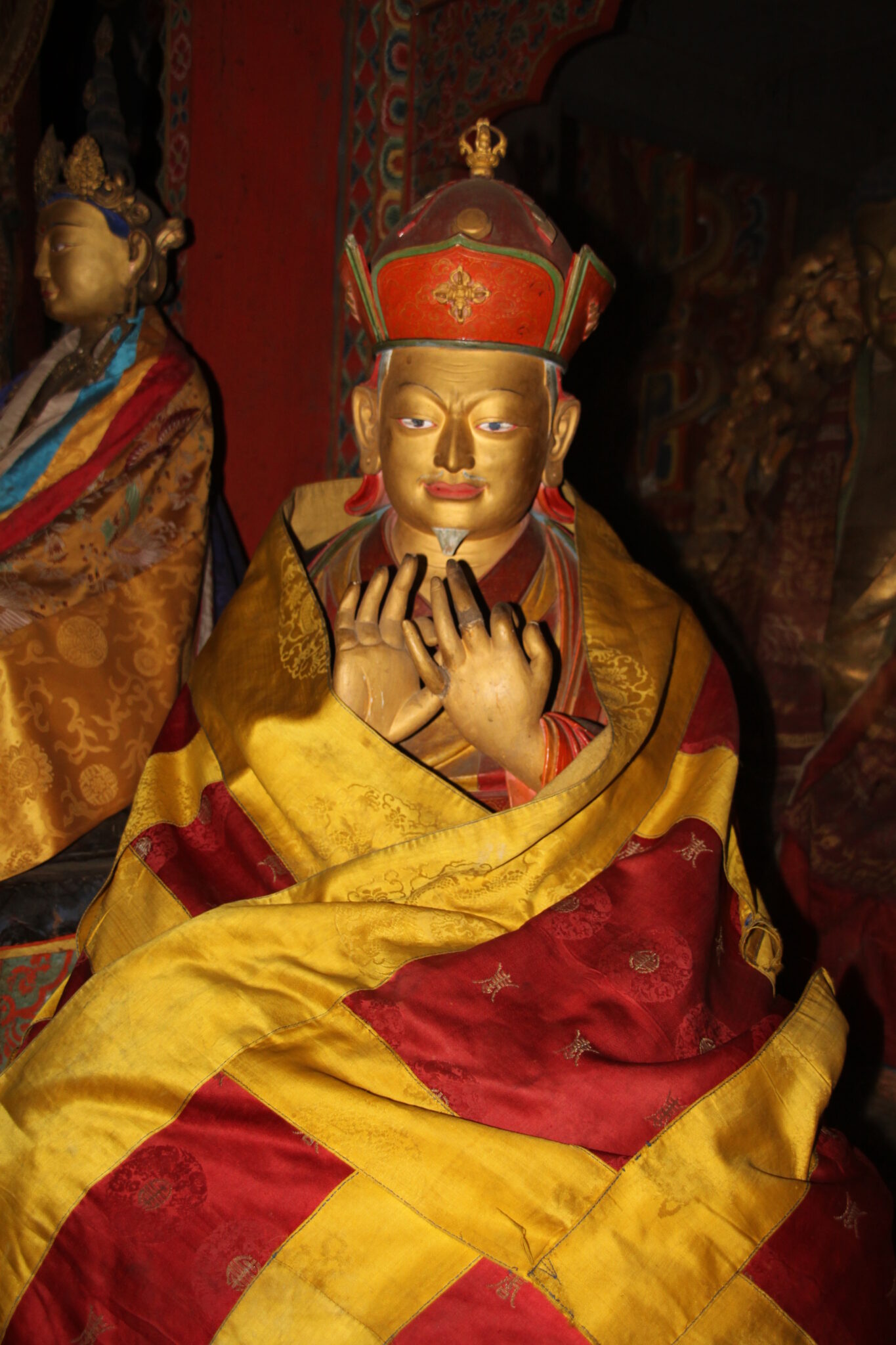 Golden statue of seated man wearing red hat with hands raised at chest; saffron and red textile wrapped around shoulders