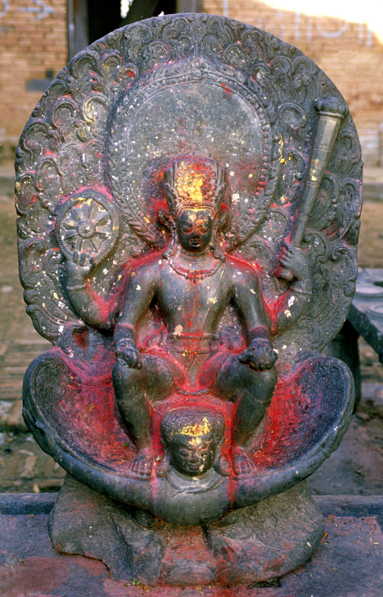 Dark stone statue of Vishnu riding Garuda; anointed with red and gold pigments