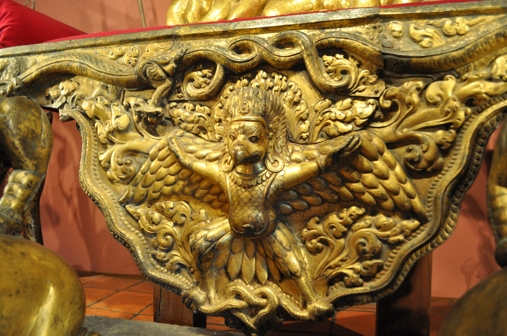 Close view of golden throne decoration depicting standing, bird-faced Garuda with outstretched arms and downturned wings