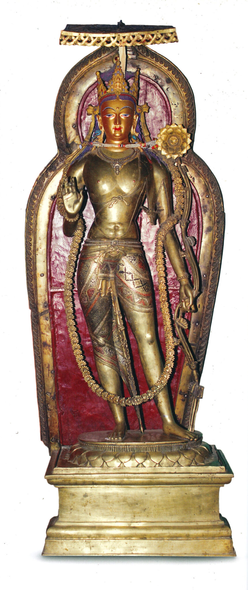 Bronze bodhisattva wearing crown, dhoti, and garland stands before red and gold nimbus; left hand turned toward viewer, right hand holds long-stemmed blossom