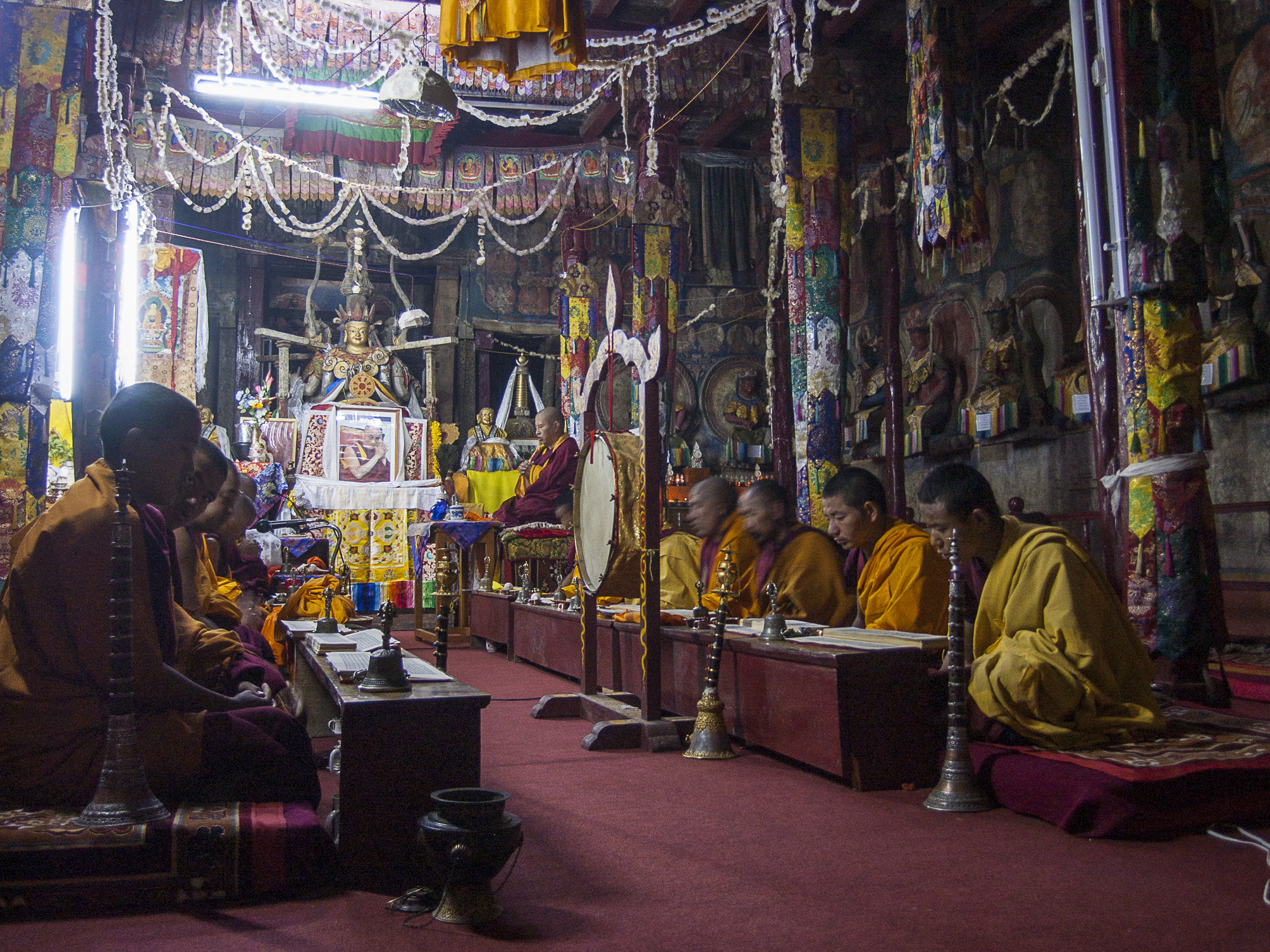 Monks sit in two rows facing one another behind short tables in hall featuring Buddha statue, banners, and decoration