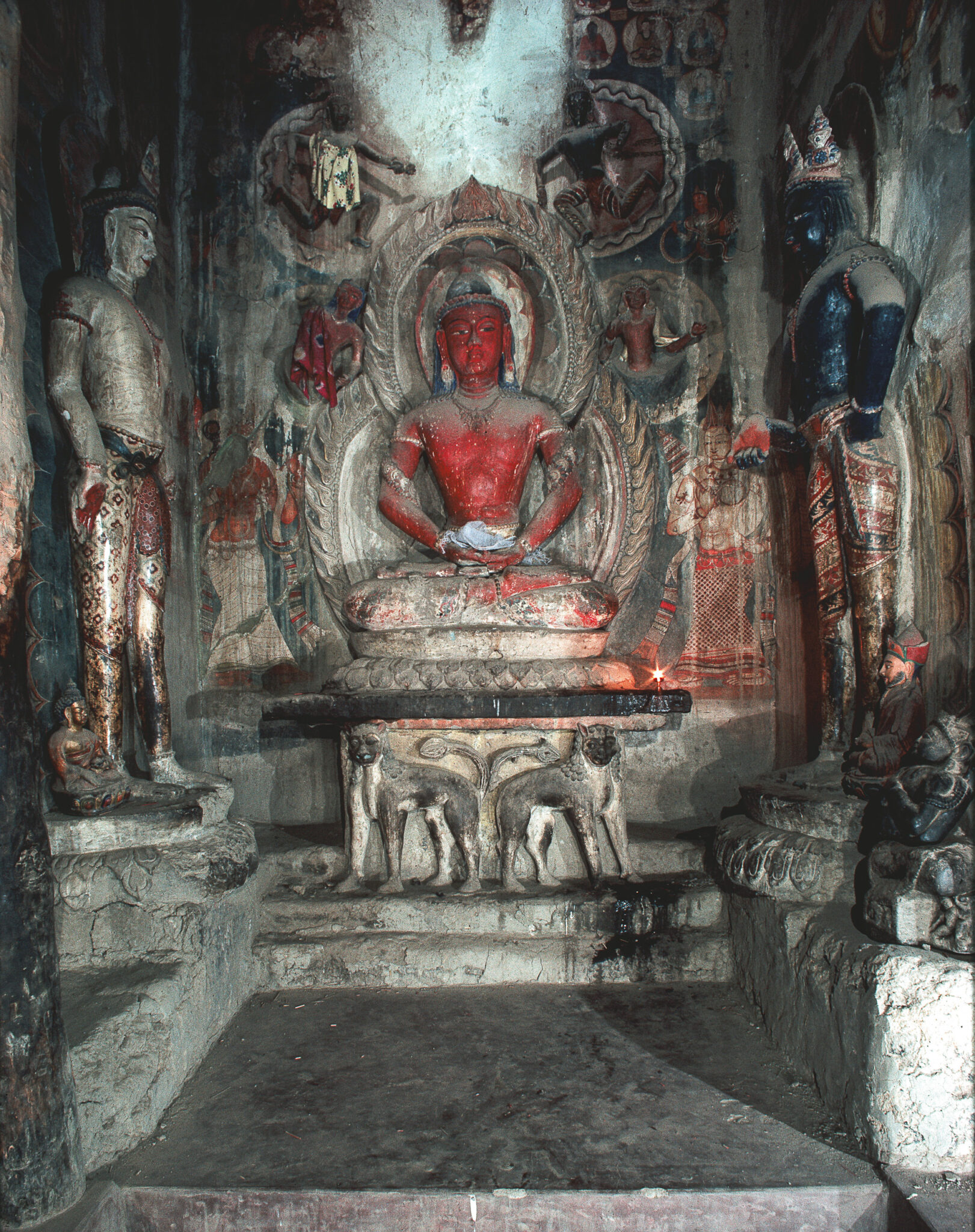 Seated Buddha statue with red skin; situated in alcove decorated with statues, reliefs, and murals depicting deities