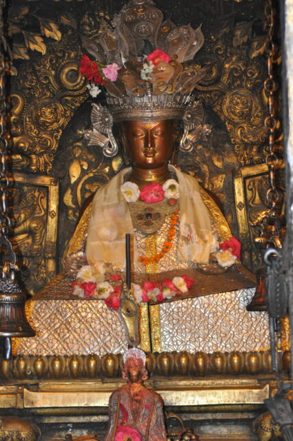 Crowned Buddha with copper face, closed eyes, and textile adornments sits before carved nimbus