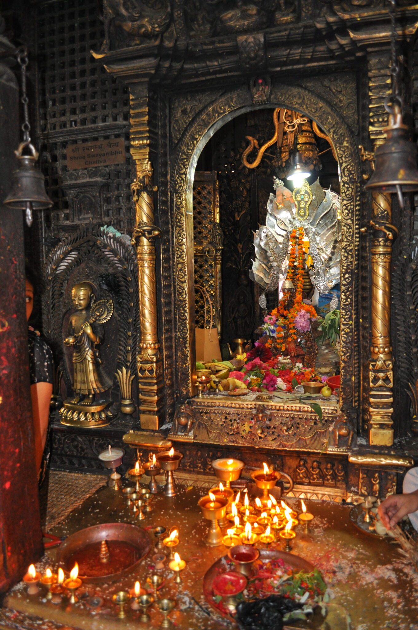 Butter lamps glow in front of columned and arched bronze niche containing devotional object adorned with garlands