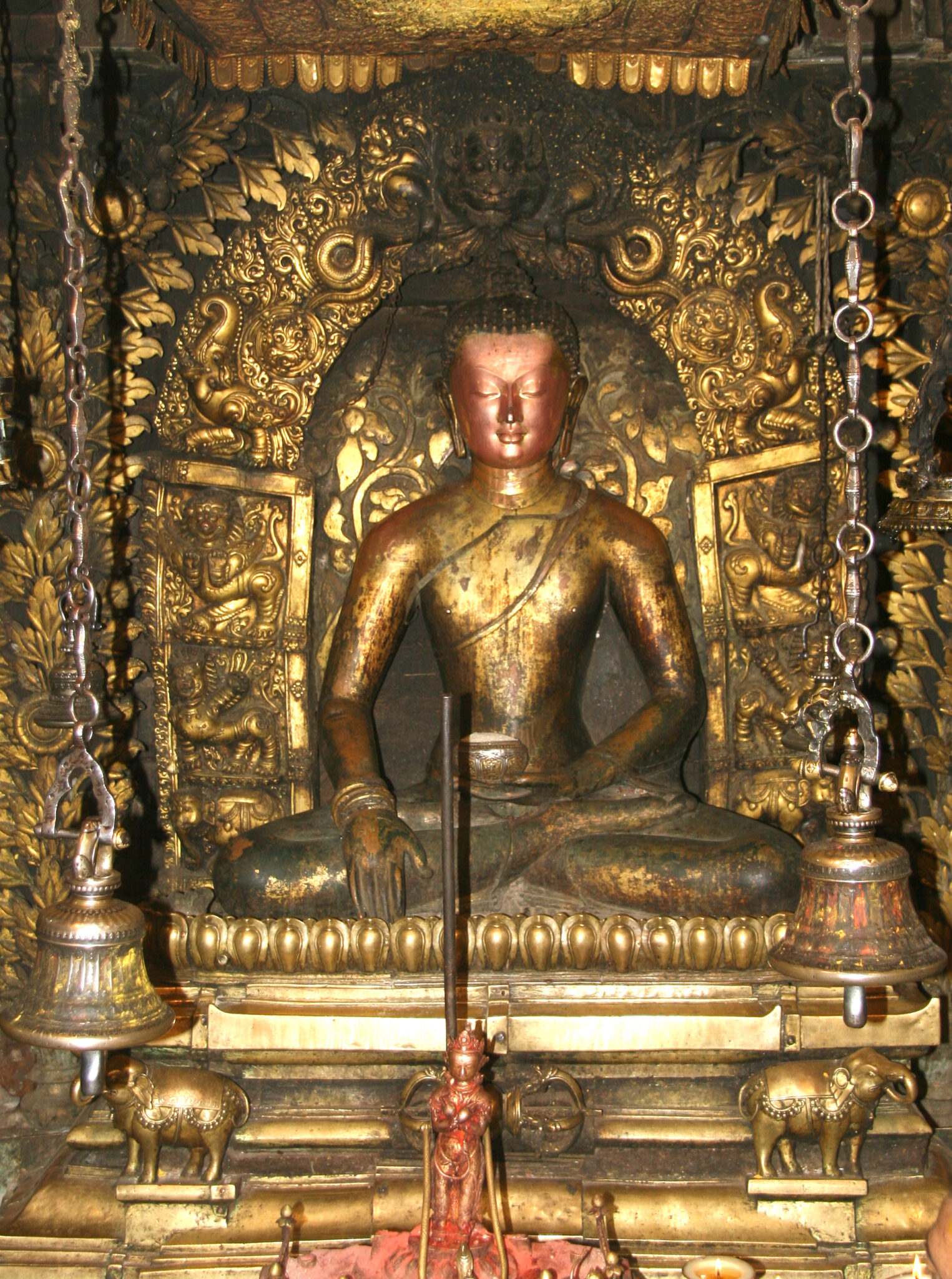 Statue of enthroned golden Buddha pictured without adornments; seated amid profusion of golden decoration and bells suspended on chains