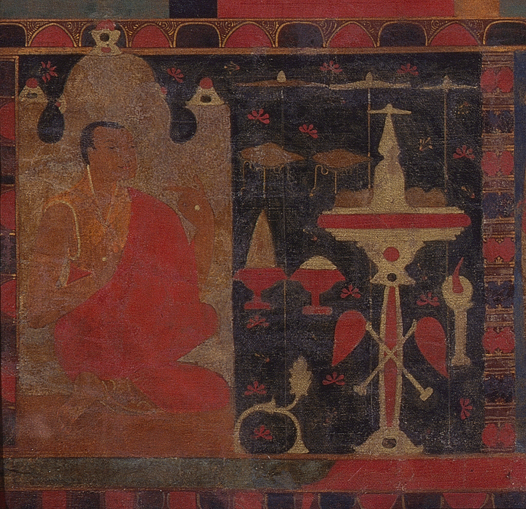 Close view of orange-robed figure seated in architectural feature on left, facing ritual objects on right