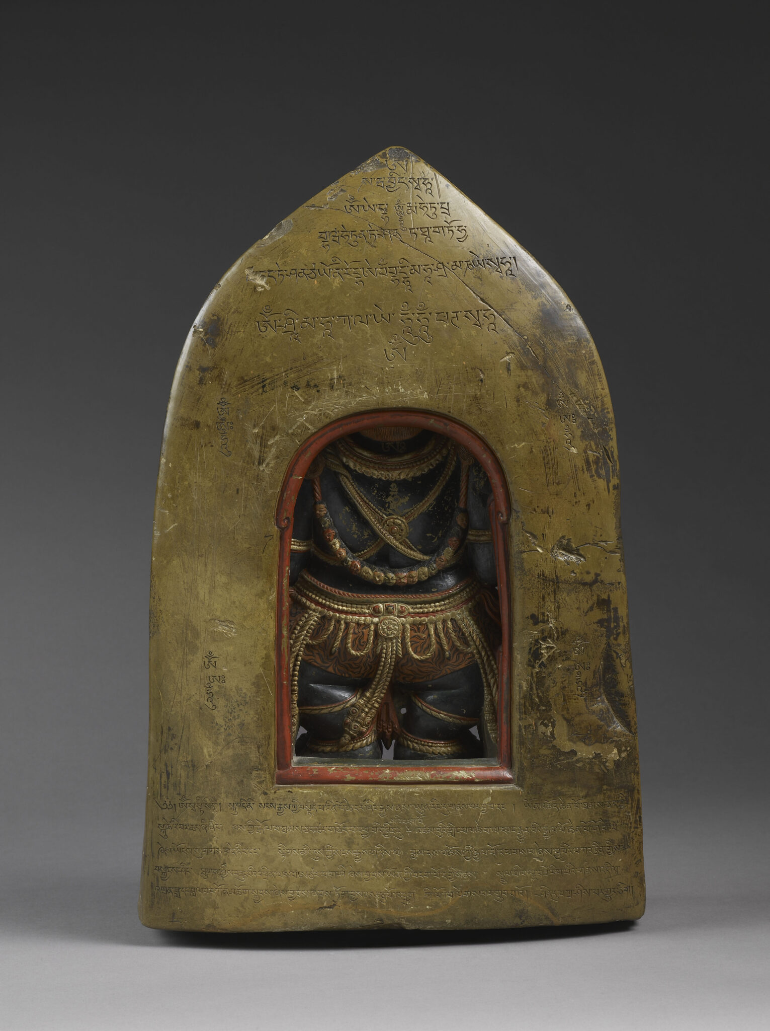 Back view of sculpture in pointed-arch shape featuring incised Tibetan script and cutout revealing backside of deity