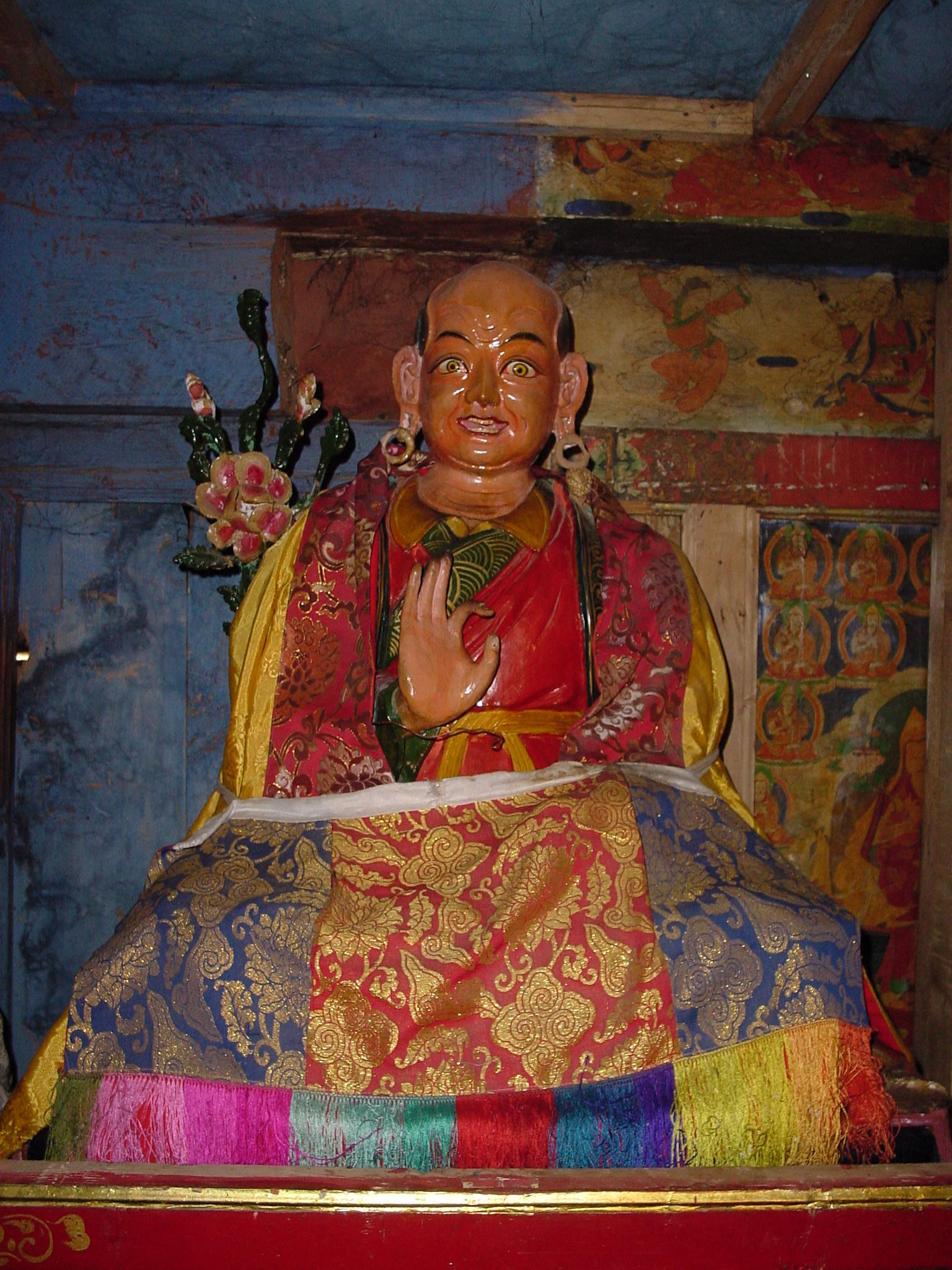 Polychrome statue of holy man holding left hand in mudra; adorned with colorful, embroidered textiles