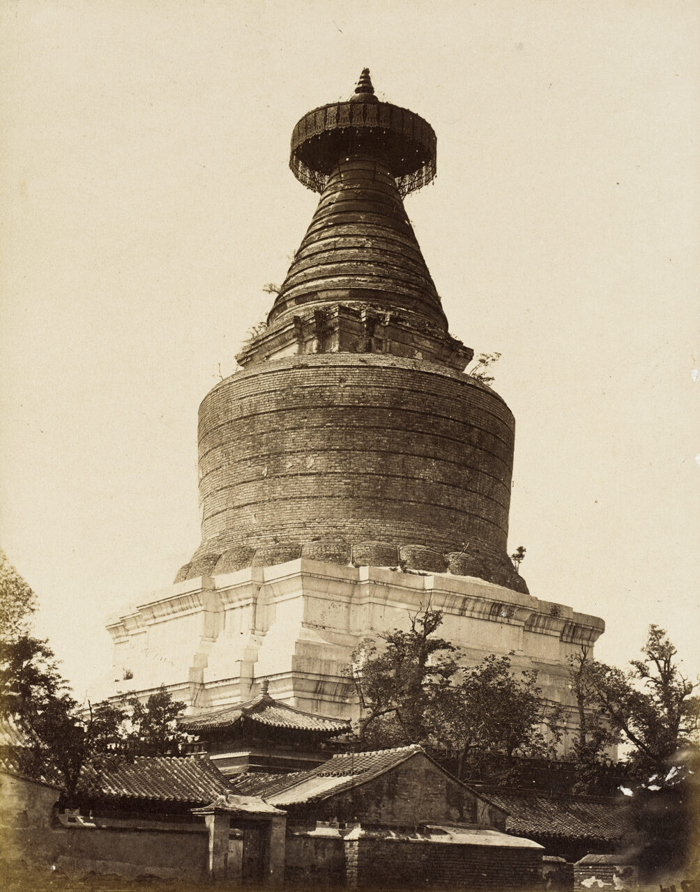Wide view sepia photograph of monumental stupa topped with canopy towering over roof and treeline