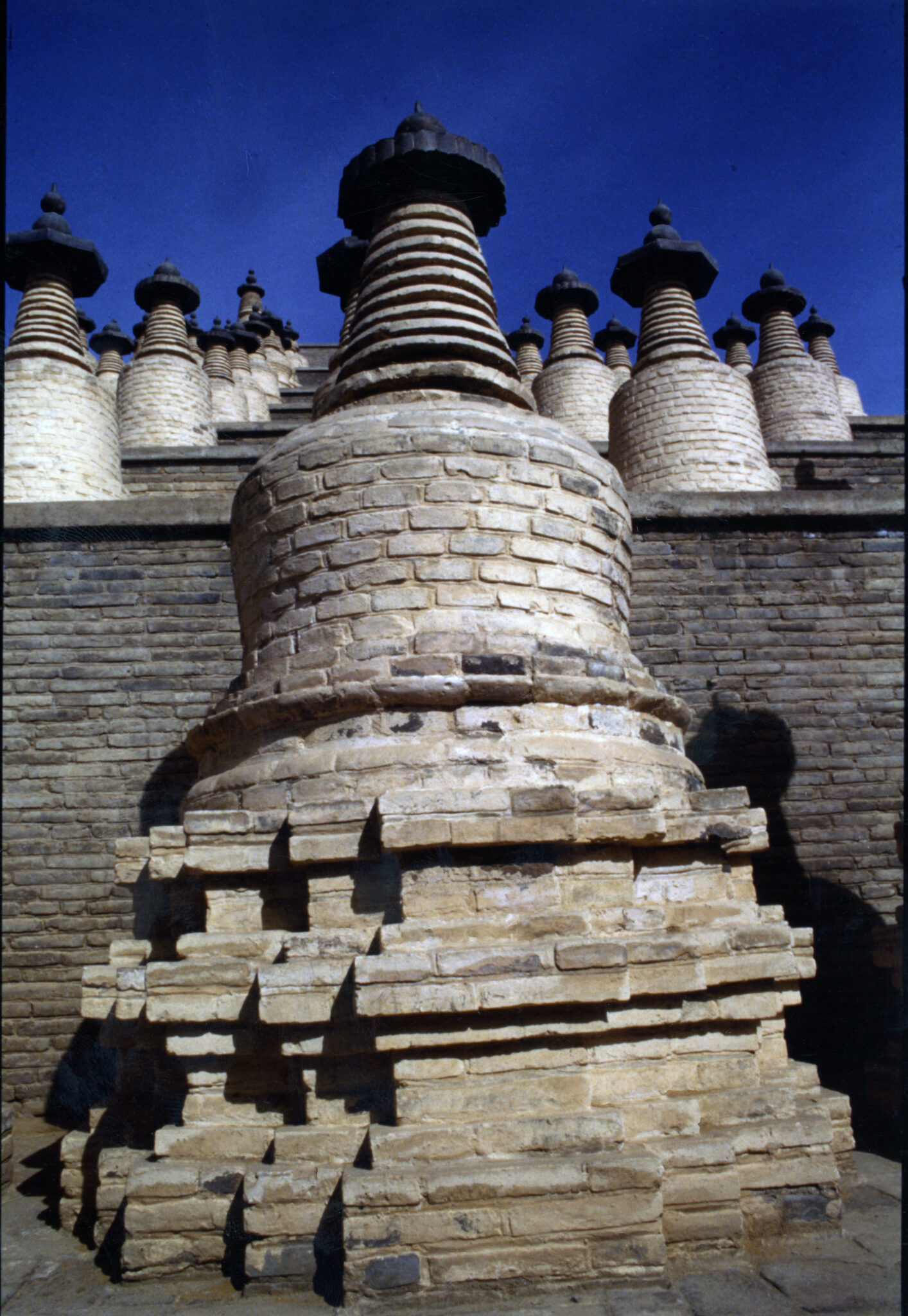 View from bottom of stupas positioned in long, ascending rows on tiers of gray brick structure