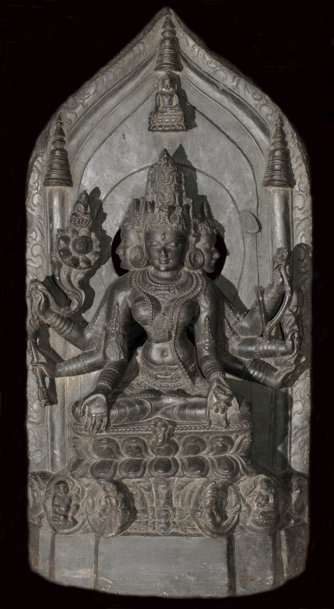 Finely carved gray relief sculpture depicting three-faced, many-armed deity holding symbolic implements before pointed nimbus