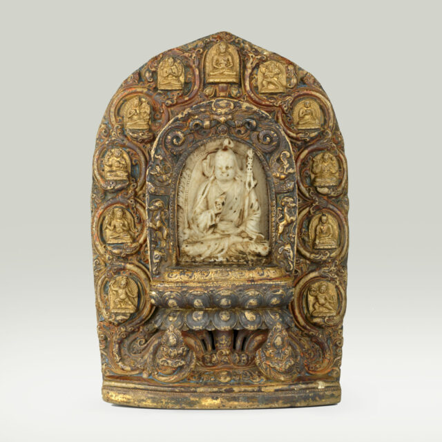 Yellow-brown stone sculpture featuring inset of holy man seated on lotus throne surrounded by nimbus of deity portraits