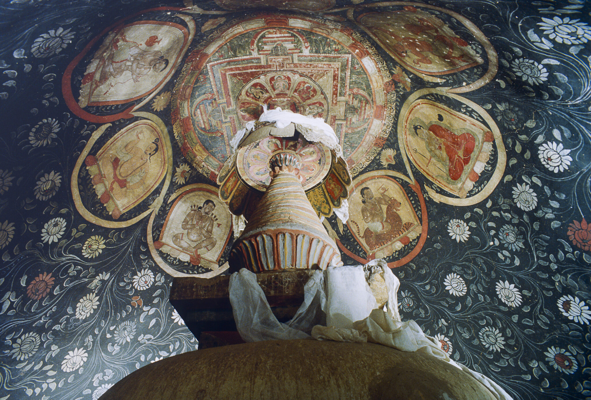 Dome viewed from base of chorten adorned with scarves; mandala at apex encircled by portraits in roundels