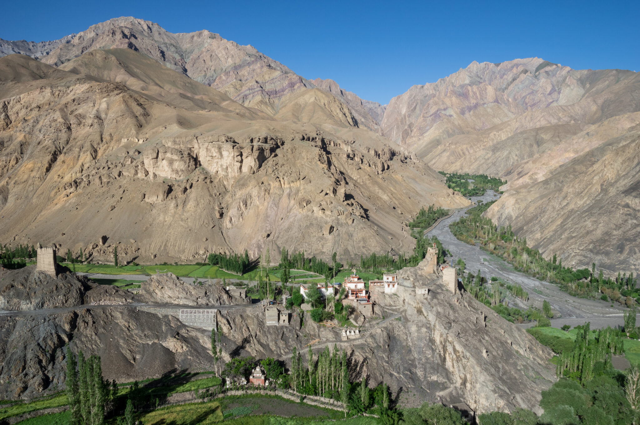 Small collection of buildings perched on outcropping situated in verdant river valley between barren mountains