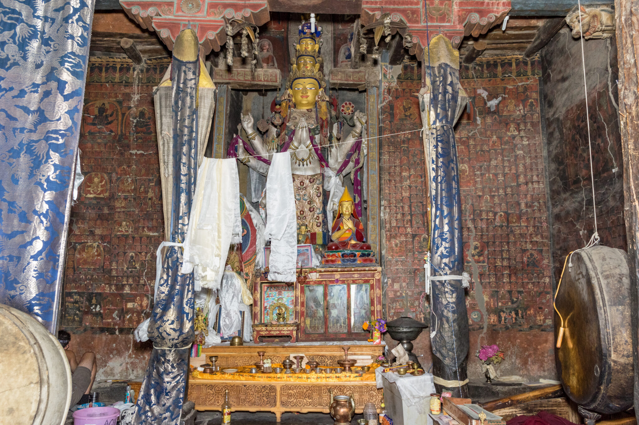 View through columns and textiles of densely muraled wall featuring niche containing tall Bodhisattva statue