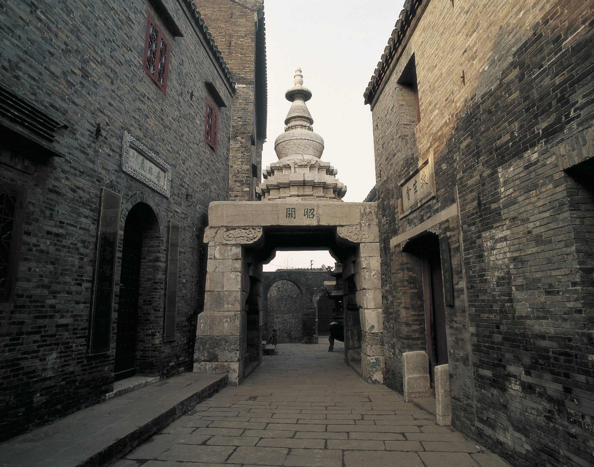 Stone gateway topped with white stupa; situated between grey-brick buildings at end of narrow street