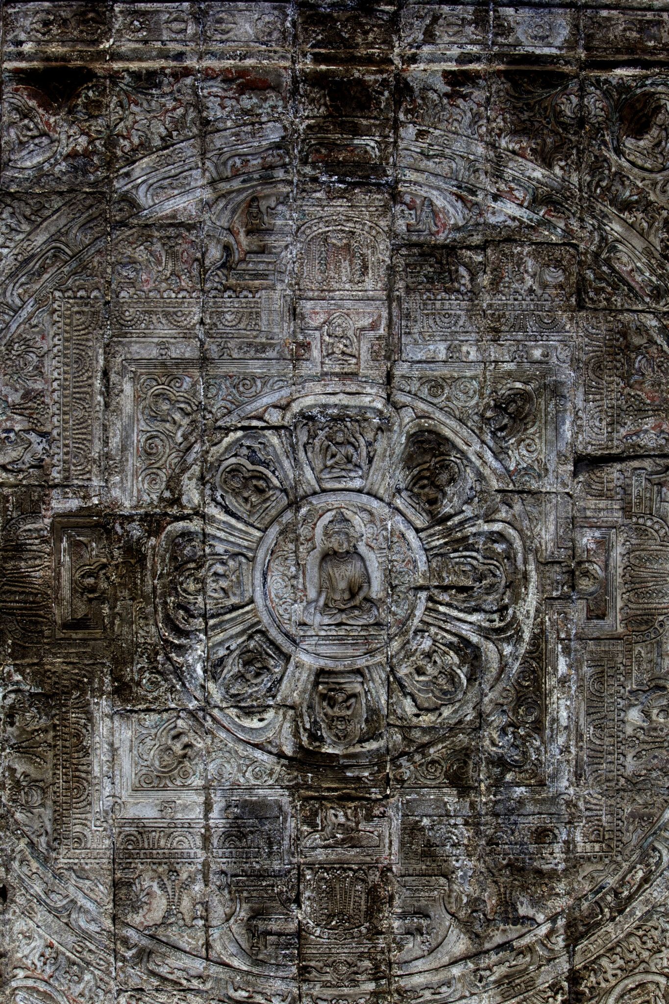View from below of weathered, mottled, densely carved stone mandala featuring seated Buddha at center