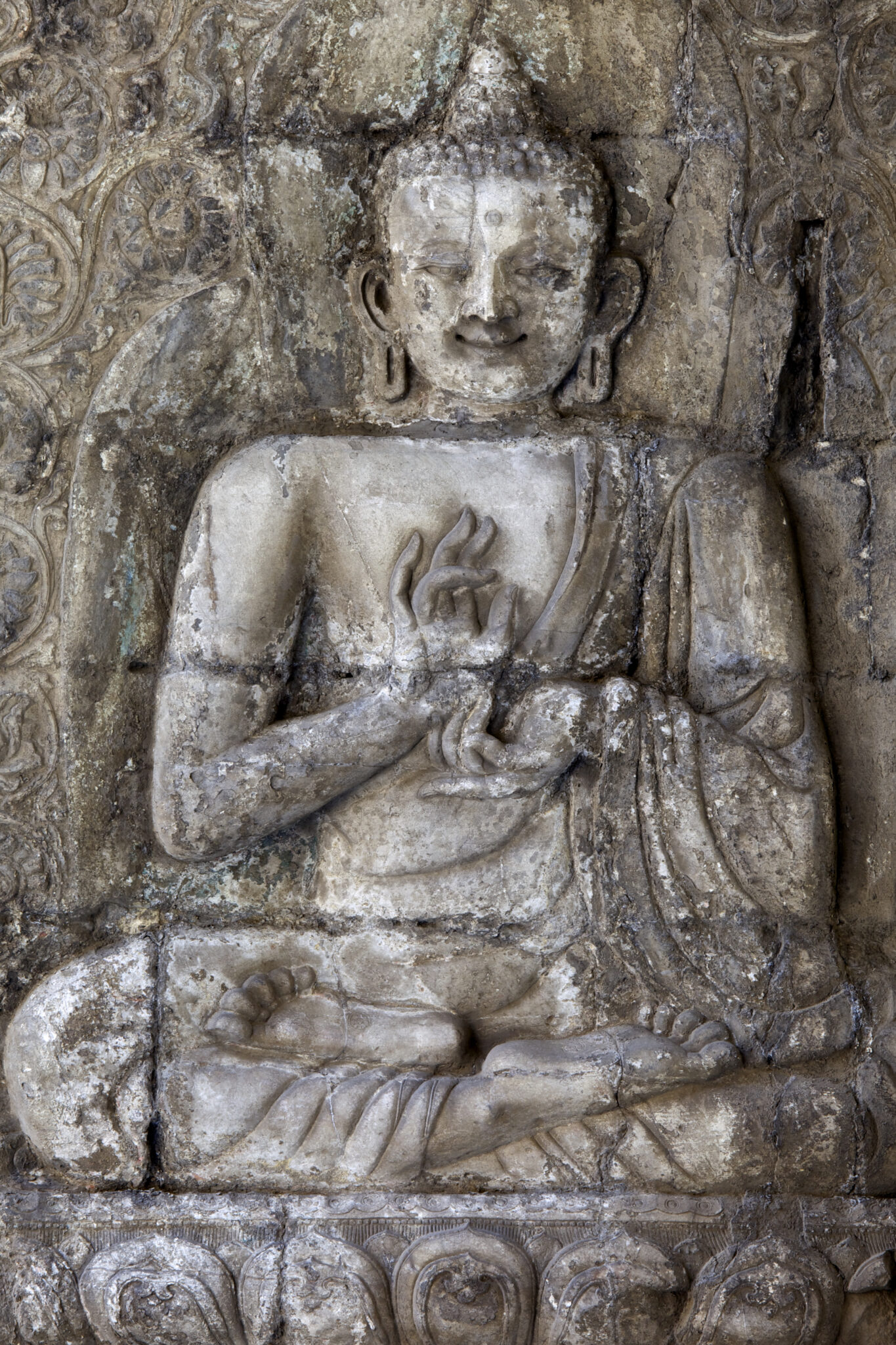 Relief on weathered stone wall depicting seated Buddha with hands posed in mudras at chest