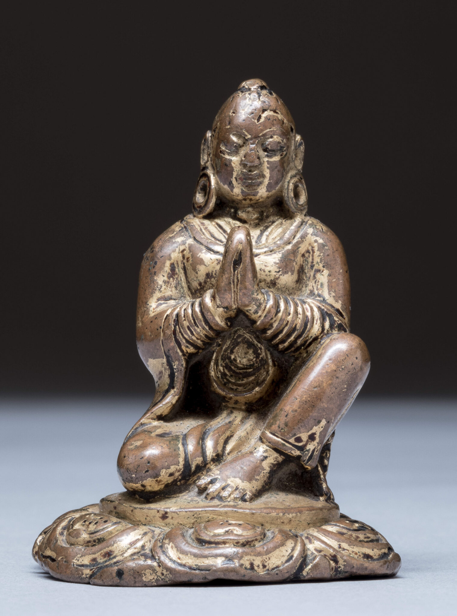 Tarnished statuette depicting figure seated with hands clasped at chest and right knee raised