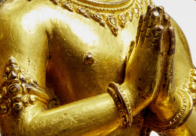 Close view of hands and chest of gilded Bodhisattva statue wearing necklace and bracelets at biceps and wrists