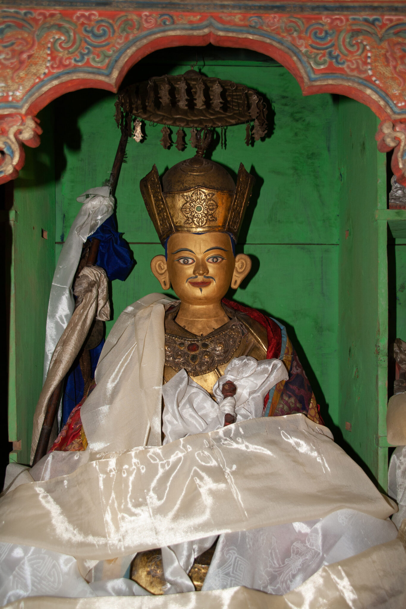 Seated statue wearing tall crown; adorned with scarves and situated in green niche behind painted red arch