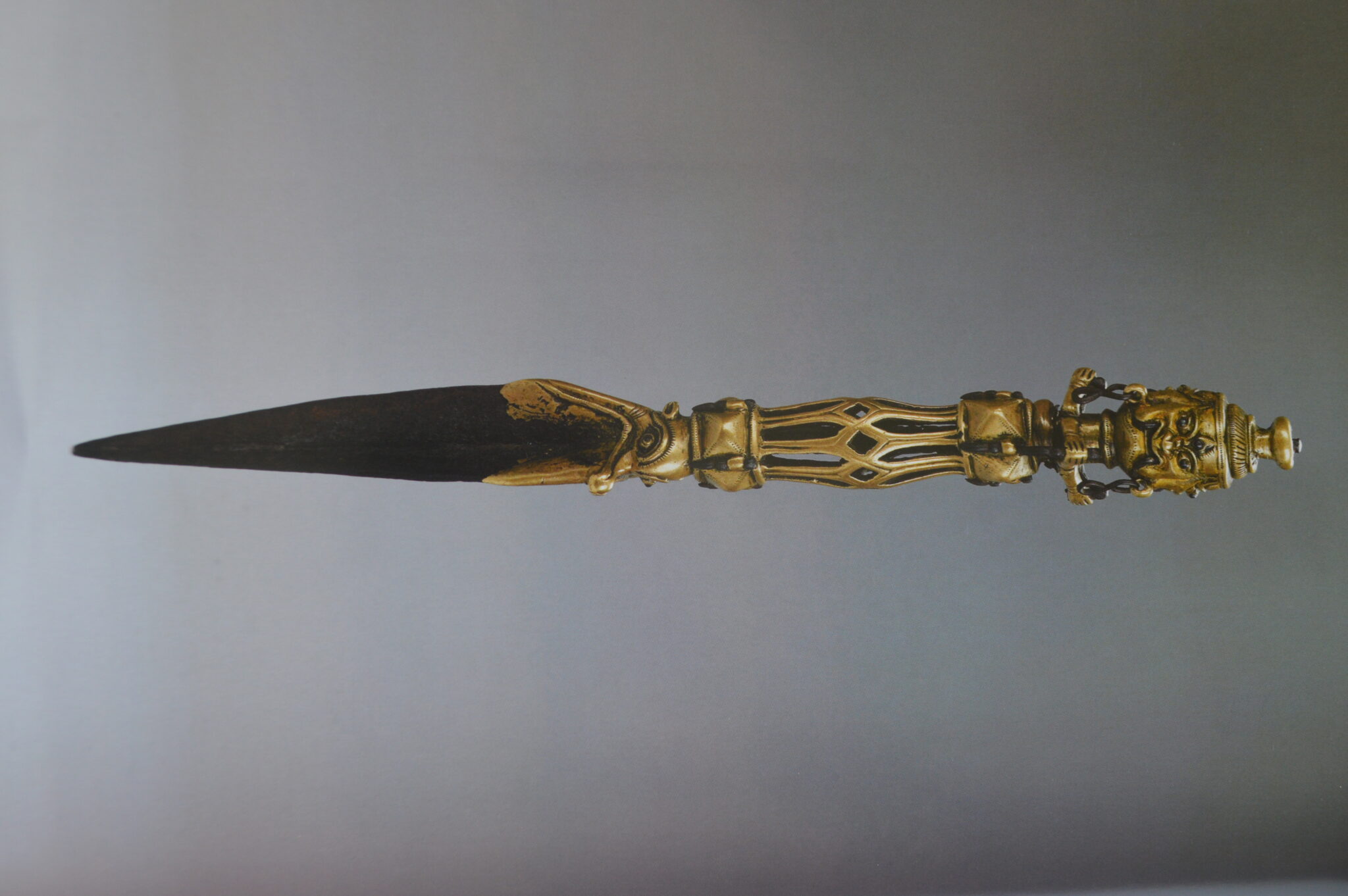 Brass-colored dagger with sharp, darkened blade; features open metalwork hilt and anthropomorphic face at pommel
