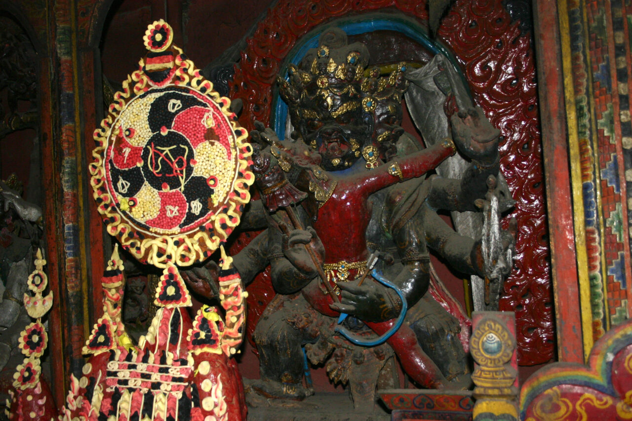 Red, yellow, and black sculpture composed of disc atop tiered structure standing before sculpture depicting two embracing deities
