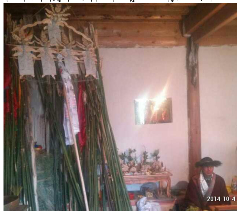 Religious implement constructed of green material, decorated with red and white scarves; situated in timber-ceilinged room