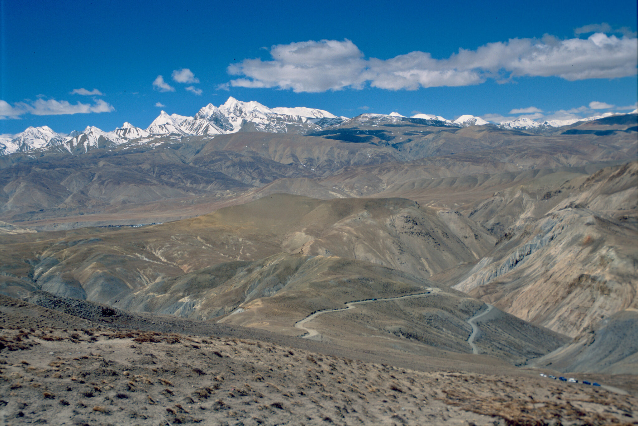 Aerial view of arid mountain foothills before range of snow-capped peaks under azure-blue sky