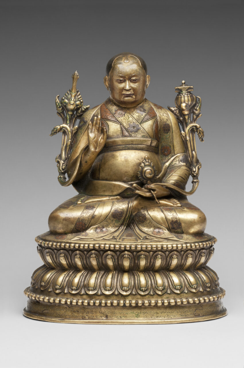 Brass-colored statue depicting figure seated on lotus pedestal holding long-stemmed blossoms in either hand