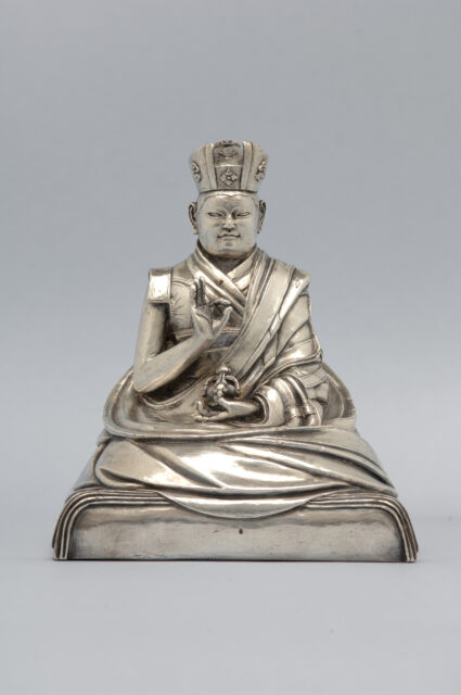 Silver statuette depicting seated figure with left hand posed in mudra, right hand holding implement