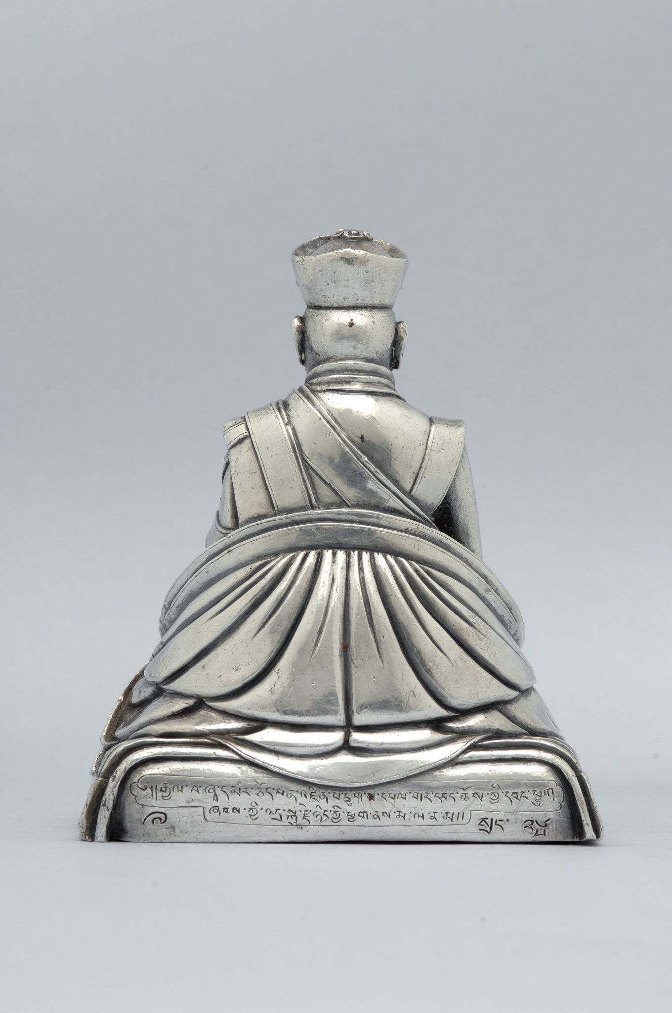 Back view of silver statuette depicting seated figure wearing finely modeled robe; two lines of text inscribed on base