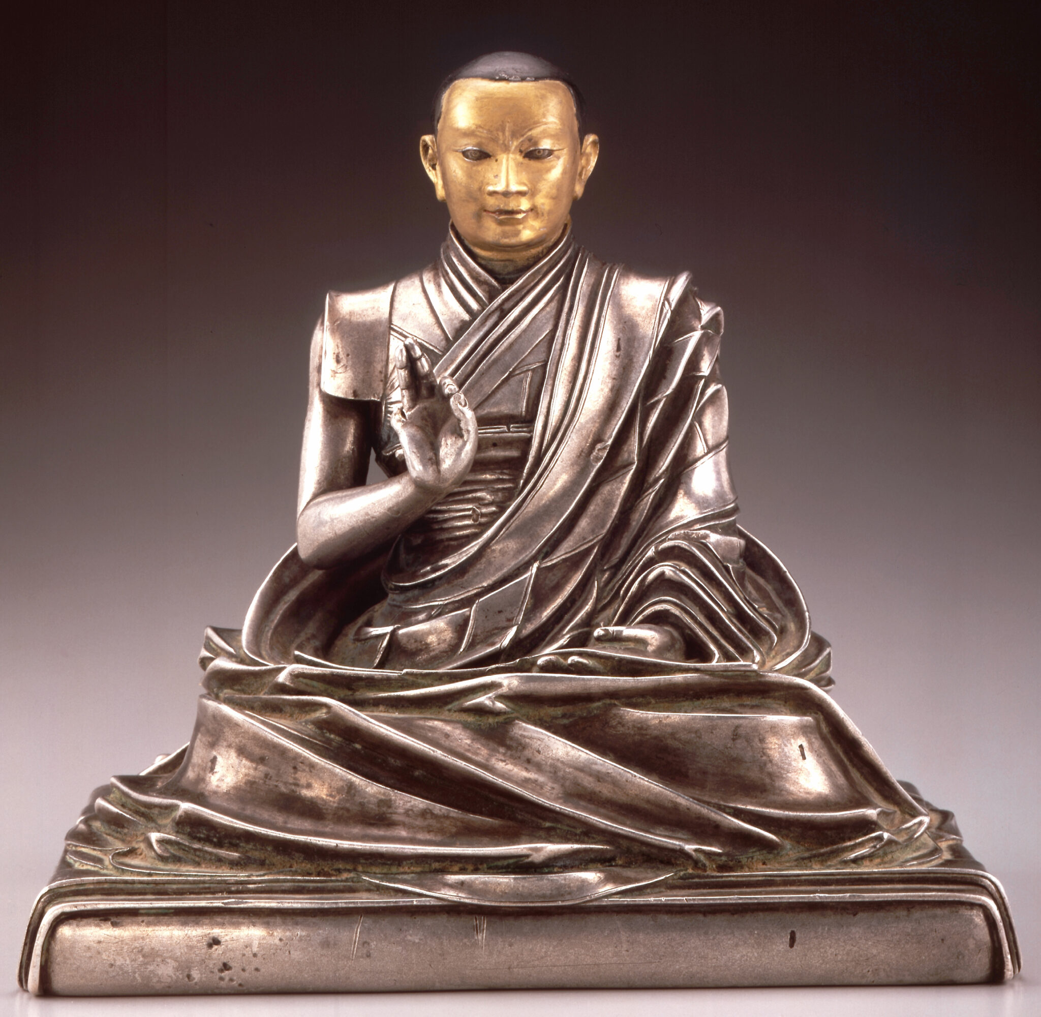 Golden-faced silver statuette depicting seated lama with left hand raised in mudra wearing finely modeled robe